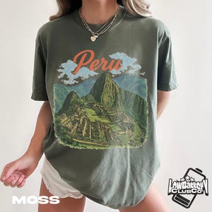 Peut inclure: T-shirt vert mousse avec un motif de Machu Picchu et le mot "Peru" en orange. Le t-shirt a un aspect vintage. Le logo "Low Battery Club Co" est visible en bas à droite.