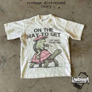 May include: A white t-shirt with a distressed vintage look. The shirt features a graphic of a frog riding a turtle with the text "On the way to get my shit together".