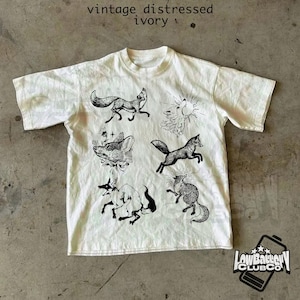 May include: Ivory-colored t-shirt with a distressed vintage look. The shirt features several black illustrations of foxes in various poses, along with the text "vintage distressed ivory". The shirt also has the logo of "Low Battery Club Co."