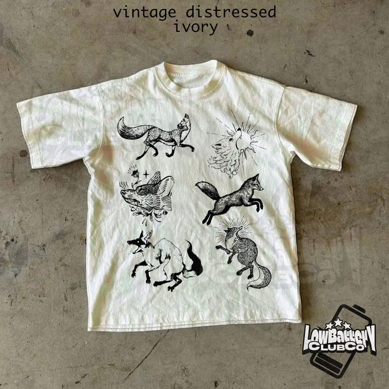 May include: Ivory-colored t-shirt with a distressed vintage look. The shirt features several black illustrations of foxes in various poses, along with the text "vintage distressed ivory". The shirt also has the logo of "Low Battery Club Co."