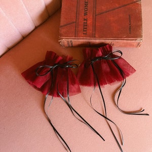 May include: Two red tulle wrist cuffs with black ribbon ties. The cuffs are lying on a pink surface with a book titled "Little Women" by Louisa May Alcott.
