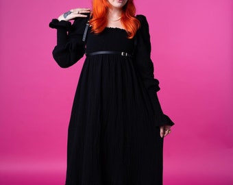 Pode incluir: Um vestido longo preto com decote quadrado e mangas compridas. O vestido tem um corpete justo e uma saia fluida com bainha com babados. Um cinto preto com detalhes dourados marca a cintura. A modelo tem cabelo laranja.