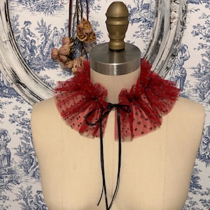 May include: A red tulle collar with black polka dots and a black satin ribbon tie. The collar is displayed on a mannequin. The background features a blue and white toile wallpaper and a decorative oval frame.