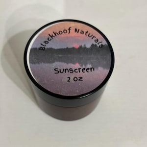 Può includere: Un piccolo barattolo rotondo di crema solare con coperchio nero. L'etichetta presenta il testo "blackhoof Naturals" sopra "Sunscreen 2 oz" e un'immagine del tramonto.