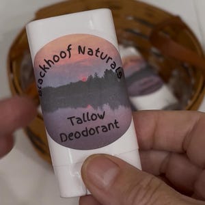 Op de afbeelding: Witte deodorantstick met een etiket met de tekst "blackhoof Naturals Tallow Deodorant". Het etiket toont een zonsondergang met een paarse lucht en boom silhouetten. De deodorant wordt in een hand gehouden.