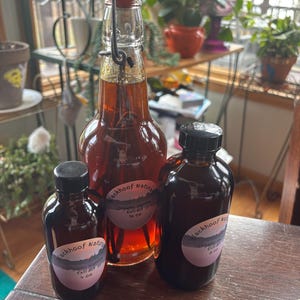 Könnte beinhalten: Drei Glasflaschen Vanilleextrakt auf einer Holzoberfläche. Die größte Flasche hat einen Bügelverschluss und enthält 473 ml. Zwei kleinere Flaschen mit schwarzen Kappen enthalten 118 ml und 237 ml. Jede Flasche trägt das Etikett "Blackhoof Naturals Organic Vanilla Extract."