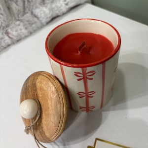 Puede incluir: Una vela roja en un recipiente de cerámica color crema con detalles de lazos rojos. La vela tiene una mecha de madera y va acompañada de una tapa de madera con un pomo blanco. Sobre una superficie blanca.