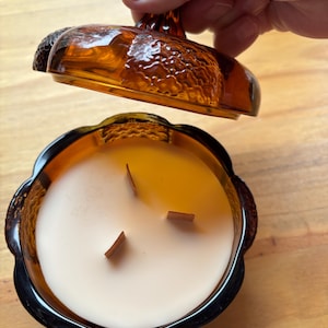 May include: A pumpkin-shaped amber glass candle jar with a matching lid held above it. The candle features three wooden wicks set in smooth, cream-colored wax. The glass container has a textured, ribbed exterior surface and a scalloped rim.