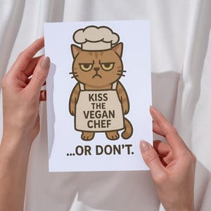 May include: A white card with a cartoon illustration of a brown cat wearing a chef's hat and apron. The apron has the text "Kiss the Vegan Chef...or don't."