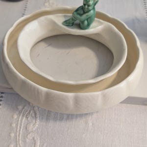 Puede incluir: Un cuenco circular de cerámica color blanco roto con borde festoneado y acabado liso. Una pequeña figura verde sentada de una criatura con orejas puntiagudas está unida al borde interior del cuenco, mirando hacia el centro del espacio circular.