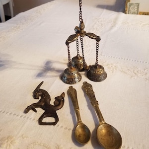 May include: An assortment of vintage brass items, including a hanging bell chime, a horse-shaped bottle opener, and two decorative spoons. The bell chime features three bells suspended by chains. The items are arranged on a white, patterned surface.