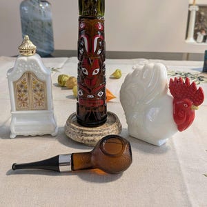 May include: A collection of decorative items including a white milk glass bottle with a gold cap, a brown glass totem pole figurine, a white glass rooster with a red comb, and a brown glass pipe with a black stem and silver band.
