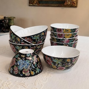 May include: A collection of ceramic bowls featuring a black exterior decorated with a vibrant floral pattern in shades of pink, yellow, blue, and green. The interior of each bowl is solid white, and the bowls are arranged in a group on a surface.