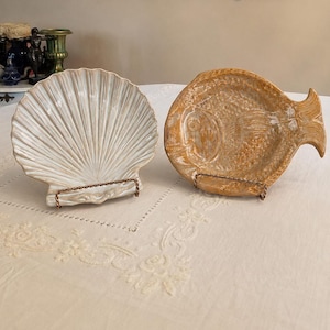 May include: Two ceramic decorative dishes displayed on wire stands. One dish is shaped like a white scallop shell with prominent ridges, while the other is shaped like a golden-brown fish with textured scales and a defined tail fin.