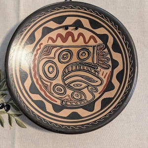Puede incluir: Un plato decorativo de cerámica redondo con base color canela y patrones circulares geométricos y abstractos pintados a mano en negro y rojo oscuro. El plato tiene un borde negro ancho con un diseño de líneas onduladas y dos orificios.