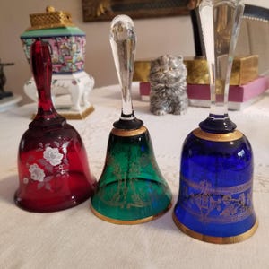 Puede incluir: Tres campanas decorativas de cristal en rojo, verde y azul. La campana roja presenta diseños florales blancos, la campana verde tiene filigranas doradas y la campana azul tiene detalles dorados. Cada campana tiene un borde dorado.