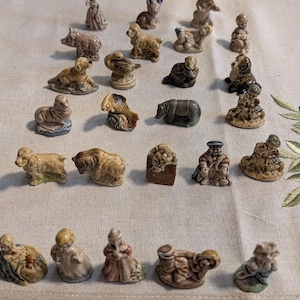 Peut inclure: Une collection de vingt-cinq petites figurines en céramique disposées en rangées, représentant divers animaux dont des chiens, un cochon, un poisson et un phoque, ainsi que plusieurs personnages humains, dans des tons terreux et brillants.