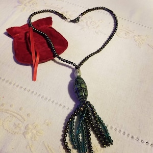 May include: A beaded necklace in a heart shape. The necklace has dark beads, a green and white pendant, and a tassel of beads. A small red velvet pouch with a red ribbon is also present.