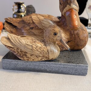 Pode incluir: Duas figuras de patos feitas de material orgânico texturizado em tons de castanho e bege, com padrões de penas detalhados e bicos lisos alaranjados. Os patos estão posicionados juntos sobre uma base retangular de pedra cinzenta salpicada.