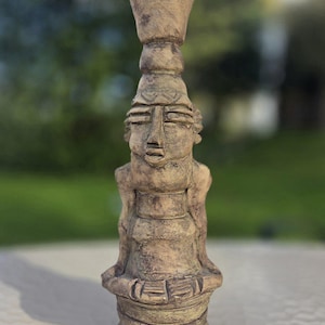 May include: A handcrafted, beige-colored ceramic candle holder in the shape of a seated figure. The figure has a detailed face, a cylindrical body, and a flared top. The sculpture is approximately 10 inches tall.