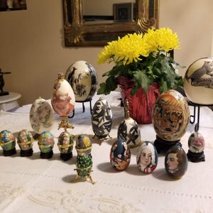 Puede incluir: Una colección de huevos decorativos, algunos sobre soportes, con diversos diseños. Los huevos presentan escenas pintadas, motivos florales y detalles intrincados. Un jarrón de flores amarillas se encuentra en el fondo, agregando color a la exhibición.