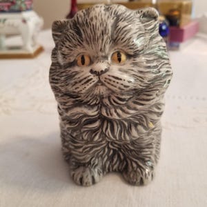 May include: A ceramic figurine of a fluffy cat with gray and white fur and yellow eyes. The cat is in a sitting position, with its paws visible. The figurine has detailed fur texture and a sweet expression.