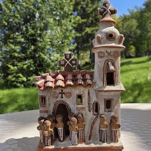 Op de afbeelding: Een gedetailleerd keramisch kerkmodel met een kruis erop en een pannendak. Het gebouw is geschilderd in tinten wit, bruin en rood, met kleine figuren van mensen ervoor. Het model staat tegen een achtergrond van groene bomen en een blauwe lucht.