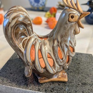 Peut inclure: Une figurine de coq en céramique avec une finition émaillée brune et crème et un design ajouré sur le corps. La sculpture présente une crête, un bec et des plumes de queue détaillés, avec un espace intérieur ouvert visible par les découpes.