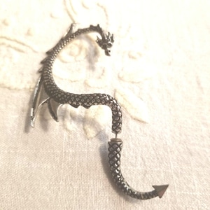 May include: A silver-toned ear cuff shaped like a dragon with a textured, scale-like body. The design features a dragon head at the top, a curved body that follows the ear contour, and a separate tail piece that attaches with a post backing.