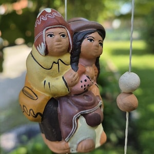 Könnte beinhalten: Ein hängendes Keramikornament mit zwei Figuren, eine trägt eine gelbe Jacke und eine gemusterte rote Mütze, die andere hat dunkles Haar in Zöpfen. Die Figuren hängen an weißen Schnüren, die mit zwei kleinen runden Perlen in Weiß und Braun verziert sind.