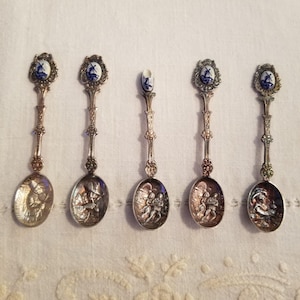 Può includere: Un set di cinque cucchiai souvenir in metallo color argento disposti in fila. Quattro cucchiai presentano terminali ovali con disegni di mulini a vento blu e bianchi, mentre il cucchiaio centrale ha un terminale a forma di zoccolo.