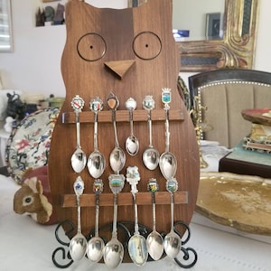May include: A wooden owl-shaped spoon rack displaying a collection of souvenir spoons. The spoons have ornate handles and small, colorful emblems. The rack is made of dark wood and has a decorative metal base.