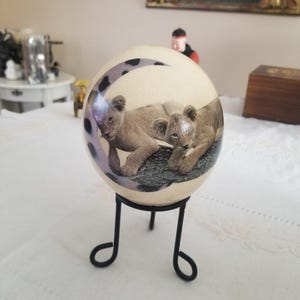 May include: A cream-coloured ostrich egg featuring a painted image of two lion cubs. The egg is displayed on a black metal stand. A crescent moon design with black spots is painted on the egg. The egg is a decorative item.