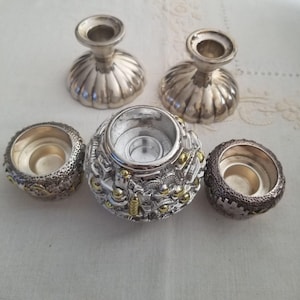 May include: A collection of silver candle holders. Two are pedestal style with a fluted design. The other three are round, with intricate detailing and gold accents. These decorative pieces are ideal for adding a touch of elegance to any space.