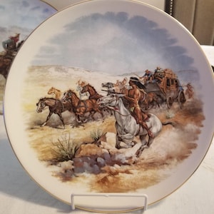Puede incluir: Un plato decorativo redondo de cerámica con borde dorado que presenta una ilustración detallada de una diligencia siendo perseguida por jinetes a través de un paisaje árido y polvoriento con vegetación escasa bajo un cielo nublado.