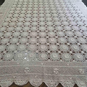 May include: Cream-colored, handmade crochet tablecloth. The intricate design features repeating circular patterns throughout the center, with a detailed border and scalloped edge. The tablecloth is laid out on a dark wooden surface.