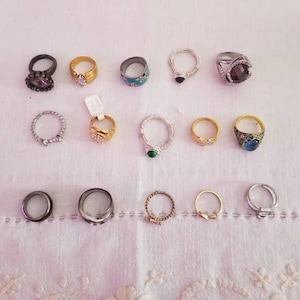 Peut inclure: Une collection de bagues variées, présentées sur une surface blanche. Les bagues présentent des designs divers, notamment des bandes dorées, argentées et noires, ornées de pierres précieuses et de détails complexes. Certaines bagues ont des pierres claires ou colorées.