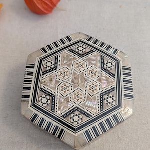 May include: A hexagonal box featuring intricate geometric marquetry with black and white striped borders. The lid displays a complex star pattern composed of inlaid mother-of-pearl, dark wood, and light wood segments, creating a detailed mosaic.