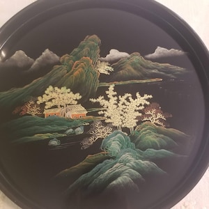 May include: A round black serving tray featuring a detailed landscape illustration in gold, green, and white. The design depicts mountains, a small house, and trees. The surface is smooth and dark, providing a high-contrast backdrop for the intricate, fine-lined artwork that covers the center of the circular tray.