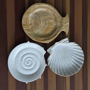 Könnte beinhalten: Ein Set aus drei Keramikschalen in Form von Meeresbewohnern. Eine Schale ist ein goldbrauner Fisch, eine ist eine weiße spiralförmige Ammonit-Schale und eine ist eine weiße fächerförmige Jakobsmuschel. Jede Schale hat eine detaillierte Textur.