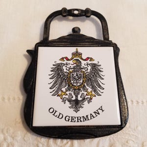 Könnte beinhalten: Ein schwarzer Metall-Topfuntersetzer mit einem weißen Kachelmittelteil mit dem deutschen Reichsadler und den Worten "OLD GERMANY". Der Topfuntersetzer hat einen Griff und eine dekorative Form.