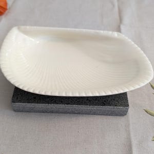 Puede incluir: Un plato de cerámica blanca con forma de concha marina, con una textura acanalada en los bordes y una superficie interior lisa. El plato descansa sobre un bloque rectangular de color gris oscuro, mostrando un diseño limpio con un borde festoneado.