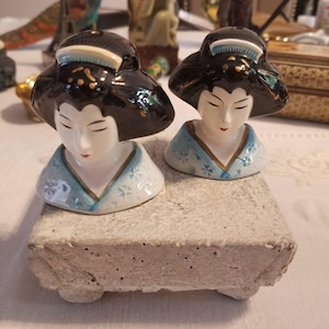 Puede incluir: Dos saleros y pimenteros de cerámica con forma de bustos de geisha con cabello negro, rostros blancos y prendas estilo kimono azul claro con pequeños patrones florales, un escote con borde dorado y un accesorio blanco para el cabello.