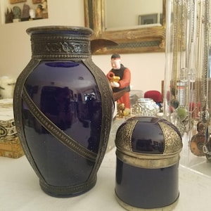 May include: A cobalt blue vase and a matching lidded jar. Both have ornate silver-toned metal trim and accents. The vase is tall and cylindrical, while the jar is round. The items are displayed on a white surface.