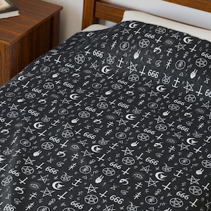 May include: Black bedspread with a repeating white pattern of occult symbols, including pentagrams, crosses, moons, and the number 666. The bedspread is on a wooden bed frame, with a wooden nightstand in the background.