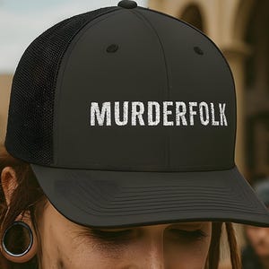 May include: Black trucker hat with the word "MURDERFOLK" in white block letters. The hat has a mesh back and a curved brim. The image shows a person wearing the hat, with dreadlocks and a nose ring.
