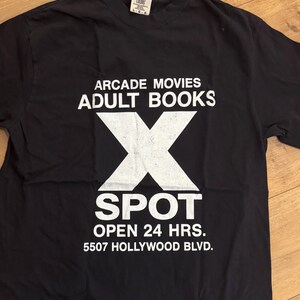 Könnte beinhalten: Schwarzes T-Shirt mit weißem Text und einer großen weißen "X" Grafik. Der Text lautet "ARCADE MOVIES ADULT BOOKS" über dem "X" und "SPOT OPEN 24 HRS. 5507 HOLLYWOOD BLVD." darunter. Das Shirt besteht aus einem weichen Material.