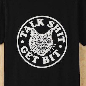May include: Black t-shirt featuring a white graphic of a cat's face within a circle. The text "TALK SHIT GET BIT" surrounds the cat. The shirt is made of a soft material and has a crew neck.