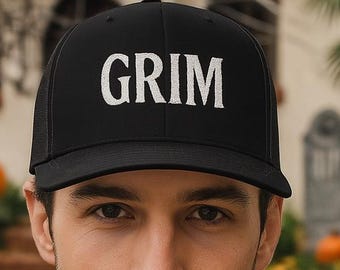 May include: Black baseball cap with the white word "GRIM" across the front. The cap is worn by a person in a black hoodie. The background includes a house with Halloween decorations and pumpkins.