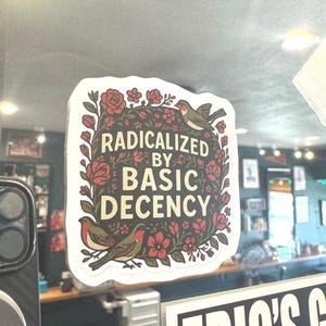 May include: A sticker with the text "RADICALIZED BY BASIC DECENCY" in a decorative font. The sticker features a floral border with red roses and two birds. The background is a light color.